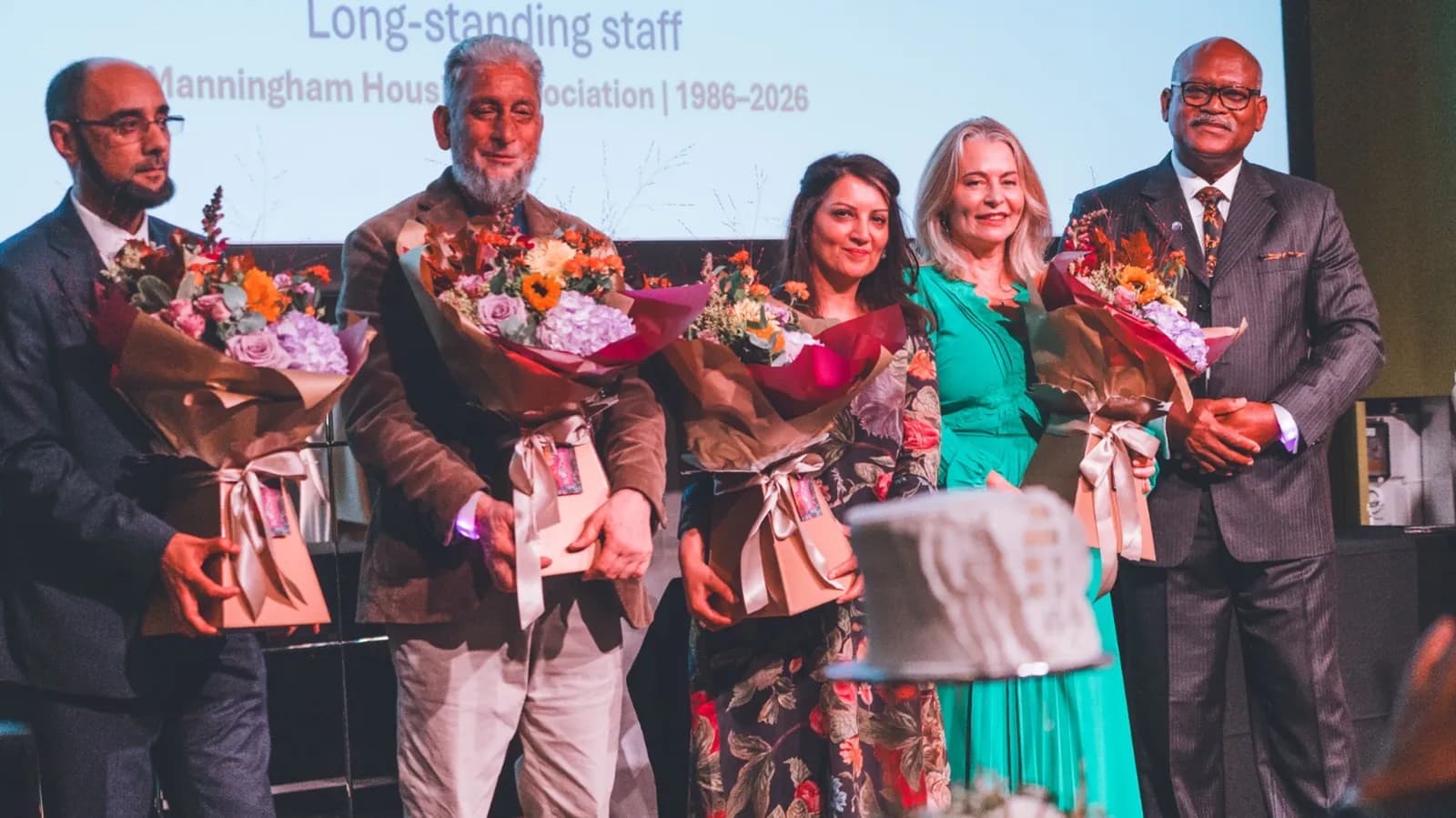 Five people, three men and two women, stand smiling and holding bouquets of flowers and gifts at an awards ceremony, with text on a screen behind them.